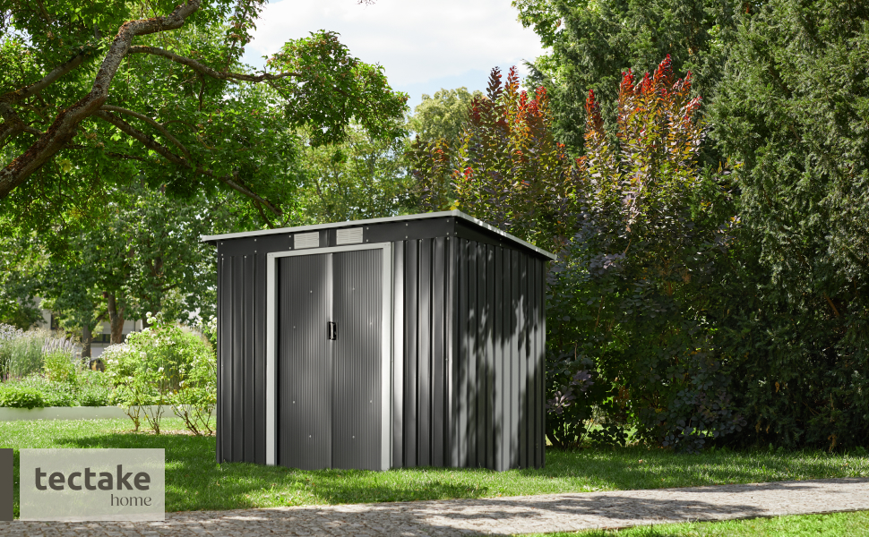 Le texte indique « tectake ». Vue extérieure d'un abri de jardin gris foncé ou d'un bâtiment de stockage avec toit en pente, positionné sur une fondation en béton dans un jardin verdoyant.