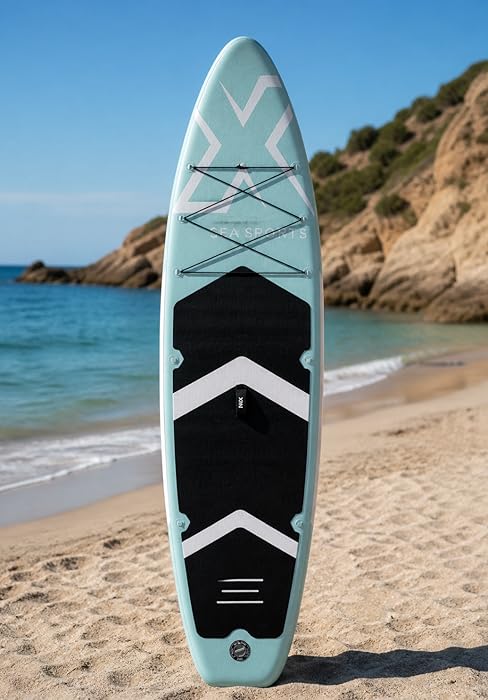 Planche de surf rayée bleu clair et noir debout sur une plage de sable. Motif en chevron à bord, positionné sur fond d'océan et de côte rocheuse sous un ciel clair.