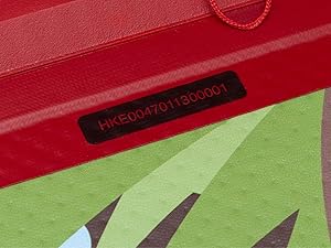 Gros plan d'une surface rouge avec une étiquette noire affichant le code « HKE004701130000001 ». Une partie d'une zone à motifs verts est visible ci-dessous.