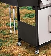 Gros plan d'un grill extérieur ou d'un chariot de cuisine avec des ustensiles en acier inoxydable suspendus à des crochets. Comprend des panneaux latéraux noirs, des roues pour la mobilité et se positionne sur de l'herbe sèche.
