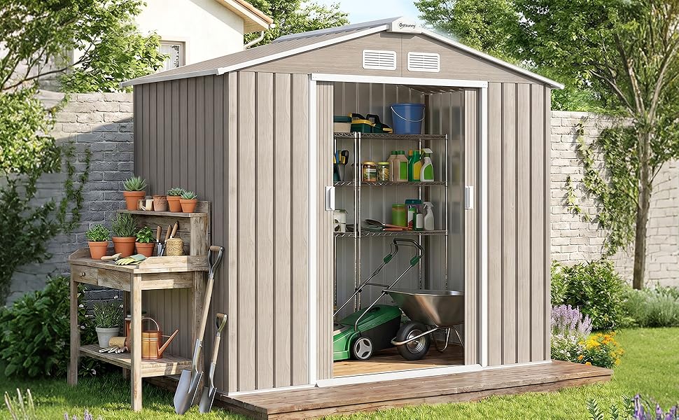 Hangar de rangement en métal avec panneaux ondulés verticaux dans la cour arrière. Abri de jardin de couleur gris/beige avec porte ouverte montrant des étagères à l'intérieur, table de rempotage en bois sur le côté gauche.
