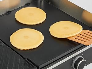 Trois crêpes dorées cuisent sur une plaque noire avec une spatule en bois visible sur le côté droit. Cadran de contrôle de température visible dans le coin inférieur.