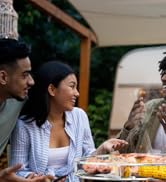 Scène de repas en plein air avec des personnes réunies autour d'une table. Produits alimentaires visibles sur la table, suggérant un repas décontracté ou un barbecue.