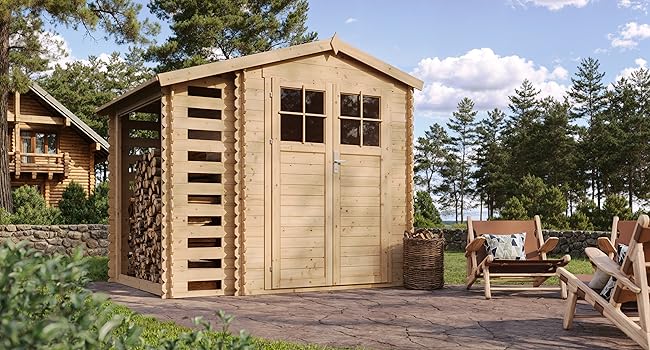 avec support de rangement pour bois de chauffage attaché. La structure présente un toit en pointe, des portes doubles avec fenêtres et est située sur un patio en pierre dans un cadre de pinède