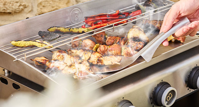 Gros plan de la viande et des légumes grillés en cours de préparation sur des grilles extérieures à l'aide de pinces, montrant le processus de cuisson.