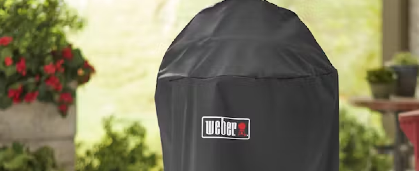 La housse de barbecue Weber noire montrée protège le gril extérieur, positionnée dans un jardin avec des fleurs rouges en arrière-plan.