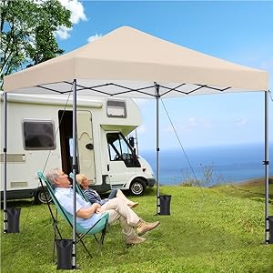 Grande tente à baldaquin beige installée à côté d'un camping-car sur l'herbe, avec une personne se relaxant sur une chaise en dessous.
