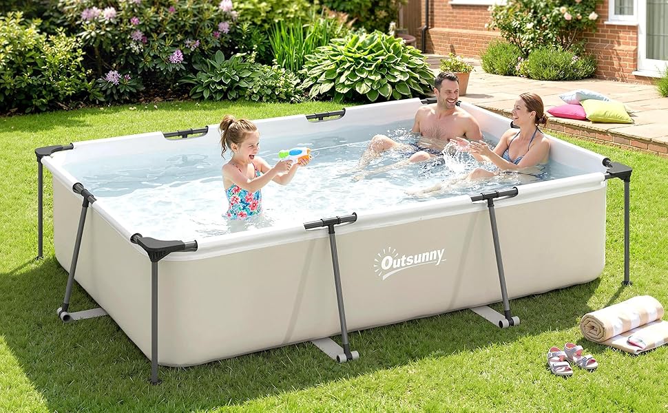 Piscine à cadre rectangulaire beige avec pieds en métal posés sur de l'herbe, contenant de l'eau avec deux enfants à l'intérieur, jouet visible sur la terrasse de la piscine.