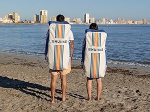 Deux personnes portant des serviettes de plage rayées avec le logo « HUIIKE » sur une plage de sable. Les serviettes présentent des rayures verticales colorées et couvrent les personnes qui les portent des épaules aux cuisses