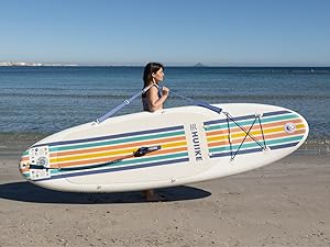 Planche à pagaie gonflable blanche avec des rayures horizontales colorées sur la plage. Comprend des cordons élastiques et une poignée de transport. Personne tenant une planche au bord de l'océan calme.