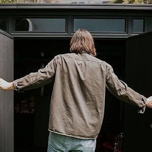 Personne portant une veste beige et un pantalon clair debout, les bras tendus, devant la porte de garage ou de grange gris foncé.