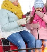 Deux personnes en vêtements d'hiver assises sur une couverture à carreaux, tenant des boissons chaudes. L'un porte un foulard jaune, l'autre un manteau et un chapeau roses.
