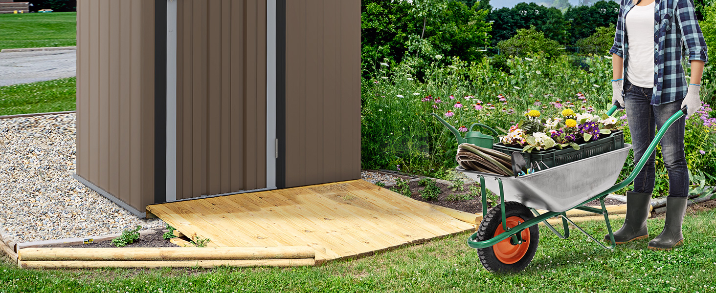 Rampe en bois menant à l'entrée du hangar métallique. Personne poussant une brouette remplie de plantes et de terre vers un jardin avec un feuillage vert en arrière-plan