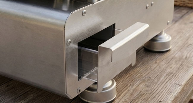 Gros plan du mécanisme du pied du meuble montrant la fixation du pied coulissant blanc sur une base cylindrique en métal, positionnée sur un fond de texture de plancher en bois démontrant la fonction de réglage.