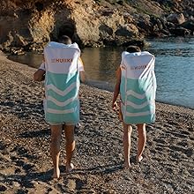 Deux personnes portant des serviettes de plage Huiike à capuche et motif à chevrons marchent sur une plage rocheuse. Les serviettes sont blanches et bleu sarcelle et couvrent les épaules jusqu'aux cuisses