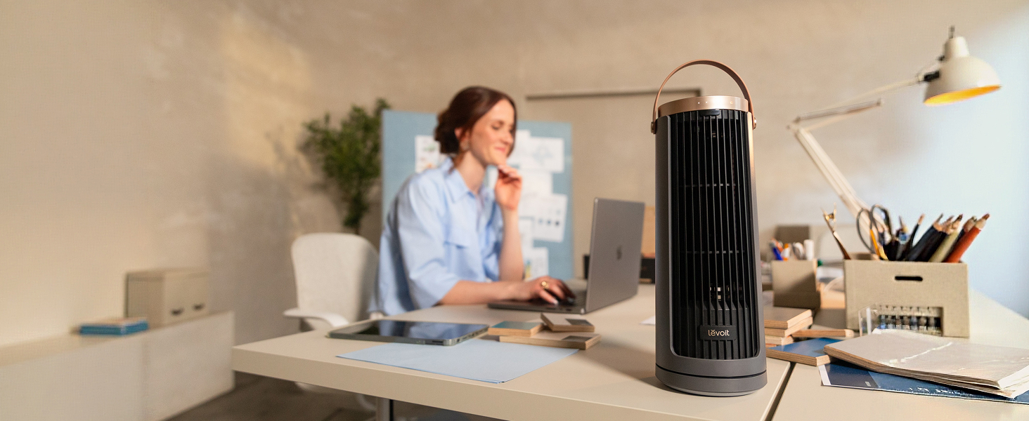 Femme assise au bureau à domicile utilisant un appareil de spa cylindrique noir. Appareil placé sur la surface du bureau à côté de l'espace de travail informatique avec des plantes et des fournitures de bureau visibles.