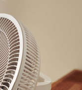 Vue rapprochée d'un ventilateur électrique blanc avec une grille métallique visible et des pales en plastique sur fond beige