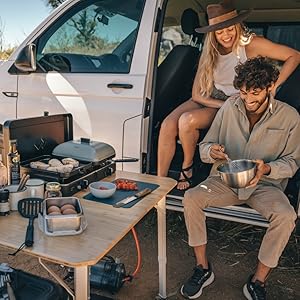 Personnes représentées assises et se prélassant dans ce qui semble être un site de camping ou de loisirs en plein air à proximité de véhicules, ce qui suggère un contexte d'utilisation mobile ou portable.