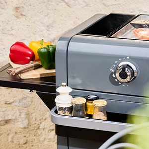 Plans rapprochés d'un grill extérieur noir ou d'un fumoir montrant un cadran de réglage de la température, une surface de cuisson et des aliments en cours de préparation.