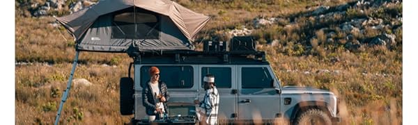 Véhicule tout-terrain avec tente de toit dans un paysage désertique, deux personnes debout à proximité, entourées d'un terrain rocheux.