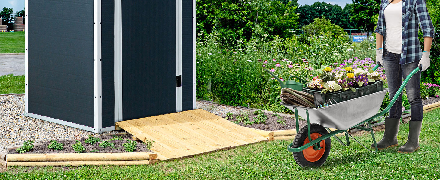 Personne poussant une brouette remplie de plantes dans un jardin. Hangar ou structure sombre visible à l'arrière-plan avec une rampe en bois.